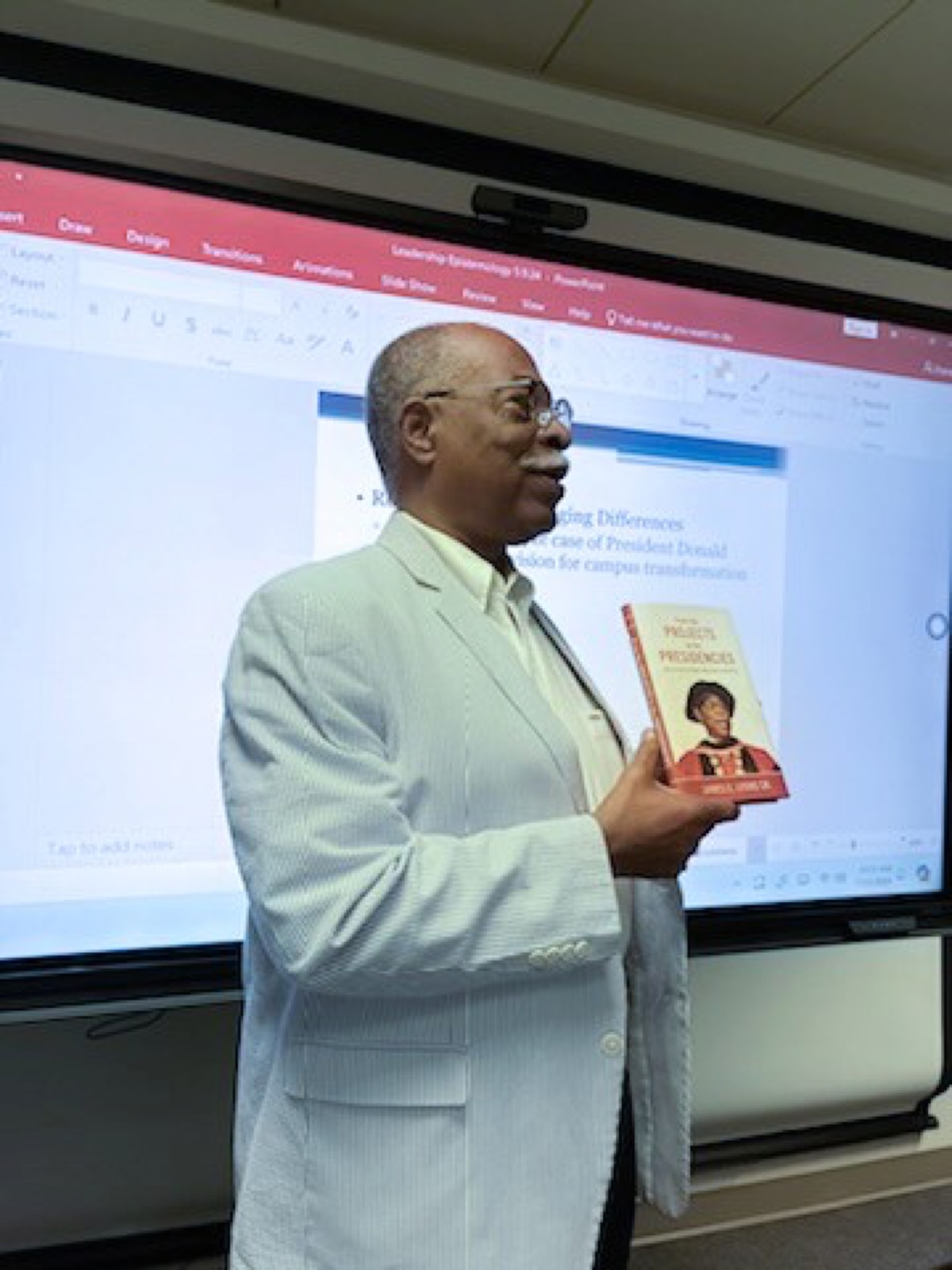 Dr. Lyons speaking to an audience while holding a copy of his memoir